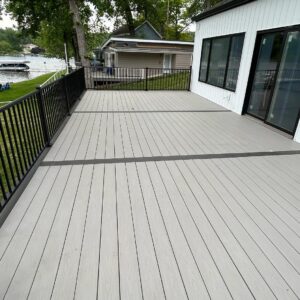 Large composite deck with black metal railings attached to a modern lakefront home, overlooking a green yard and water.