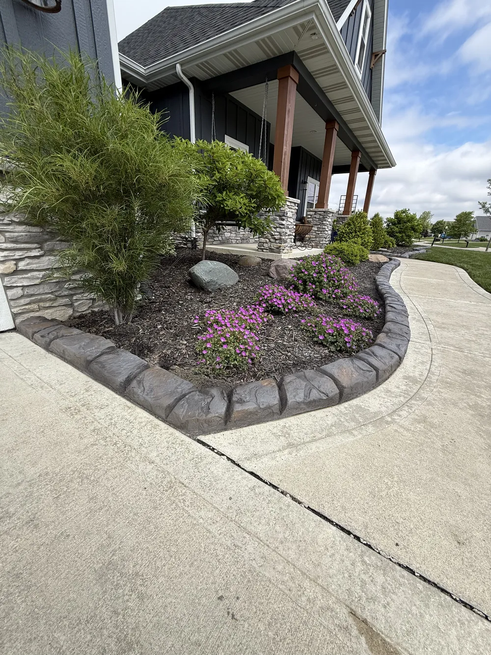 Concrete curbing outlines a front yard garden bed with shrubs, flowers, rocks, and a curved sidewalk.