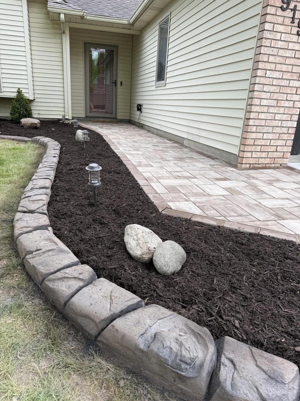 Concrete curbing frames a mulched planting bed beside a paver walkway leading to a front door.
