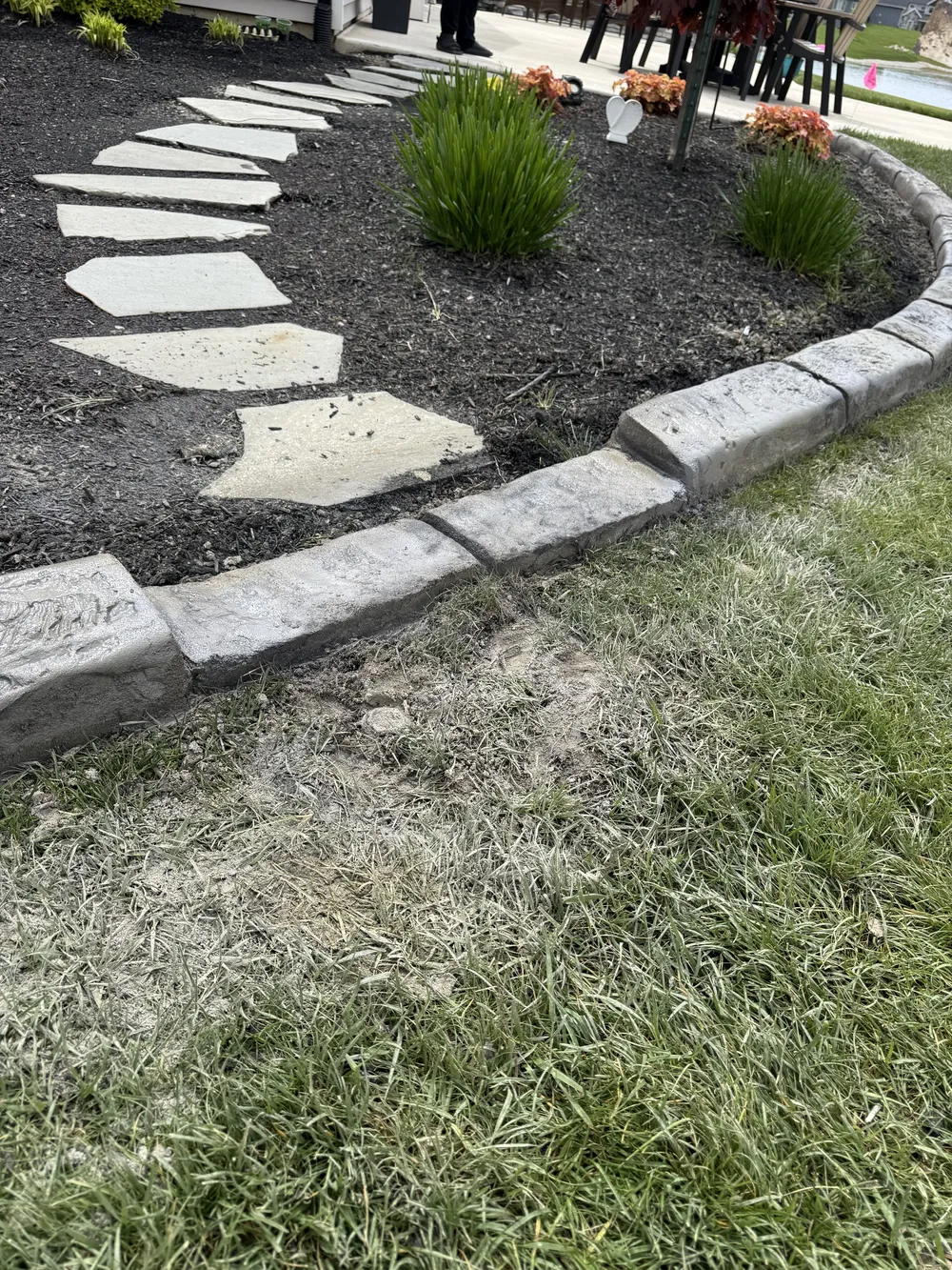 Concrete curbing edges a black mulch garden with a stone path and small shrubs beside a lawn.
