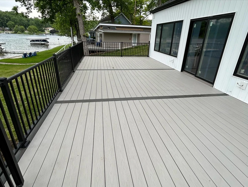 Curbscapes Outdoors deck construction huntertown in - Curbscapes Outdoors Wide light gray deck with black railings sits beside a white lake house with water and docks to the left.