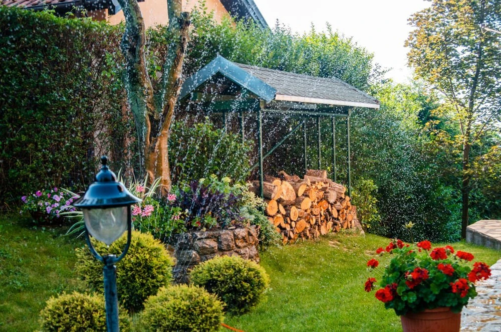 Irrigation installation mists a colorful garden with red flowers and trimmed shrubs. A covered woodpile sits behind.