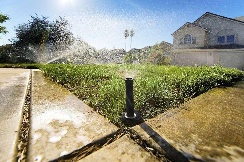 Irrigation installation sprays a sunlit front lawn between wet concrete paths and a cream stucco home.