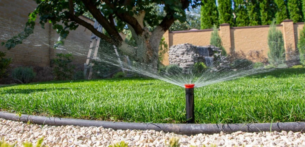 Irrigation installation fans water across vivid grass beneath a shade tree with a rock waterfall behind it.